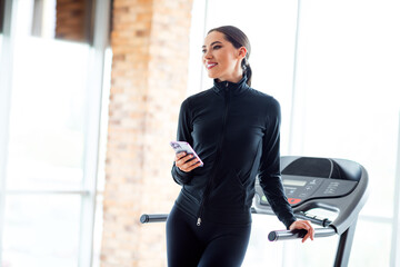 Young sportswoman using smartphone during workout on treadmill in stylish indoor gym setting with...