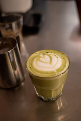 Hot matcha latte with heart-shaped latte art served in a clear glass on a stainless cafe counter. Warm cozy coffee shop mood with copy space and soft bokeh.
