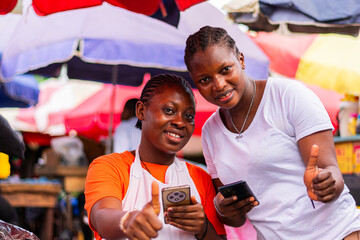 Young, excited African market woman and customer holding smartphones, giving thumbs up