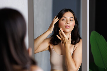 Smiling woman with perfect makeup wearing tank top looking in mirror standing at home.