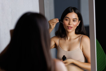 Beautiful young woman applying cosmetic serum onto her face near mirror in bathroom