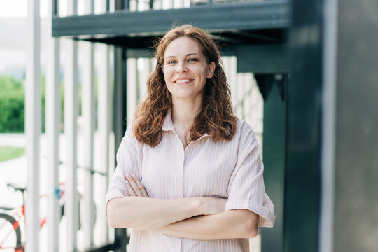 Portrait of smiling european business woman standing arms crossed out of office in front of modern building. - Powered by Adobe