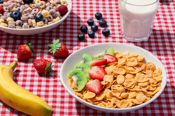Two bowls of cereal with fruits on red checkered tablecloth, one with cornflakes, strawberries, kiwi, and another with colorful loops, blueberries, raspberries, blackberry, banana, and milk