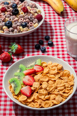 Bowl of cornflakes with fresh strawberries and kiwi on red checkered tablecloth, served alongside colorful fruit cereal, blueberries, bananas, strawberries, and a glass of milk