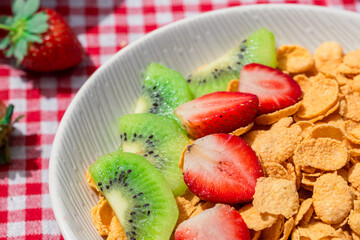 Close up of cornflakes served with fresh sliced strawberries and kiwi on red checkered tablecloth, showing healthy breakfast concept with vibrant colors, texture, and natural detail