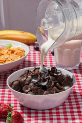 Milk pouring into bowl of chocolate cereal on red checkered tablecloth, with cornflakes, banana, strawberries, and blueberries in background, showing sweet breakfast and lifestyle nutrition