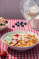 Milk pouring into bowl of cornflakes with strawberries and kiwi on red checkered tablecloth,...