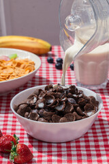 Milk pouring into bowl of chocolate cereal on red checkered tablecloth, with cornflakes, banana, strawberries, and blueberries in background, showing sweet breakfast and lifestyle nutrition