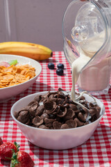Milk pouring into bowl of chocolate cereal on red checkered tablecloth, with cornflakes, banana, strawberries, and blueberries in background, showing sweet breakfast and lifestyle nutrition