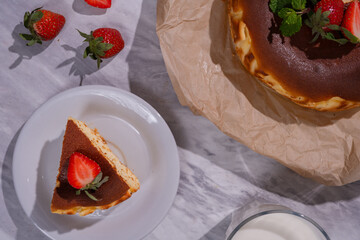 Top view of baked cheesecake with strawberries and mint on parchment paper, served with a slice on white plate, fresh strawberries, and a glass of milk on marble background