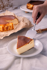 Slice of baked cheesecake on white plate with whole cake in background, placed on marble table with cloth, showing creamy dessert texture and homemade sweet pastry detail