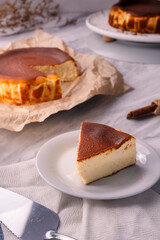 Slice of baked cheesecake on white plate with whole cake in background, placed on marble table with cloth, showing creamy dessert texture and homemade sweet pastry detail