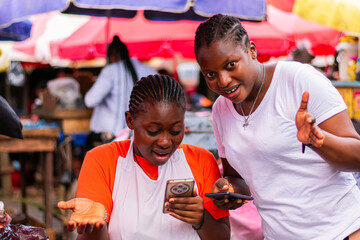Young, happy African market woman and her customer holding smartphones, overly excited