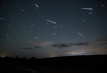 A dramatic long exposure nightscape capturing linear star trails streaking across a dark, cloudy sky above a faintly illuminated horizon, showcasing Earth's spin.
