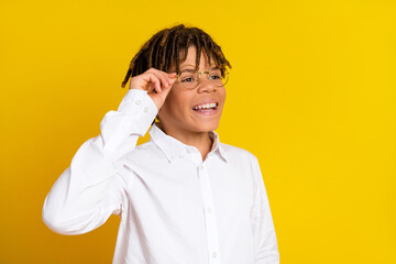 Smiling young student adjusting glasses in front of a vibrant yellow background wearing a white...