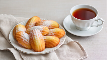A plate of golden, freshly baked madeleine cookies, lightly dusted with powdered sugar, sits on a linen cloth. A cup of hot tea next to the plate suggests a cozy and relaxing break.