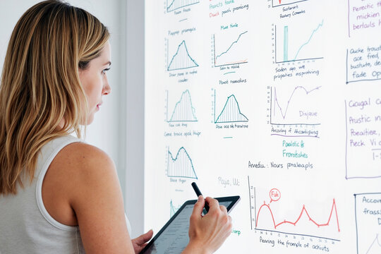 Caucasian young adult woman analyzing data charts on whiteboard while holding digital tablet, studying graphs and handwritten notes, focusing on business analytics and research