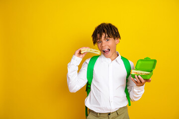 Schoolboy wearing a green backpack enthusiastically eating a sandwich from a lunchbox against a...