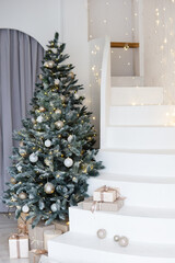 A large Christmas tree in the living room, decorated with garlands and light balloons. Bright Christmas interior with a white staircase.