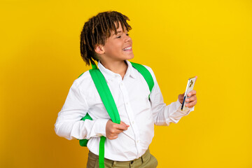 Cheerful schoolboy with green backpack smiling while holding smartphone against vibrant yellow...