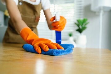A woman wearing gloves wipes and disinfects a table with spray and cloth indoors, focusing on...