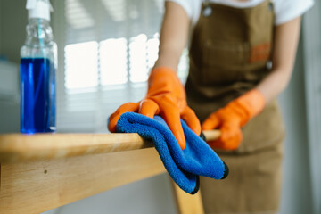 A woman wearing gloves wipes and disinfects a table with spray and cloth indoors, focusing on...
