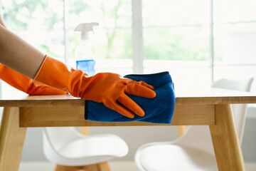 A woman wearing gloves wipes and disinfects a table with spray and cloth indoors, focusing on...