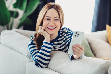 Charming woman with a happy smile relaxes indoors holding a smartphone sitting on a cozy couch in a...