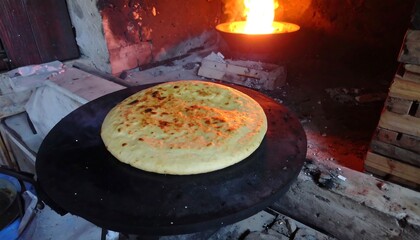 A round flatbread is cooked on a hot, dark surface, surrounded by a rustic outdoor oven.