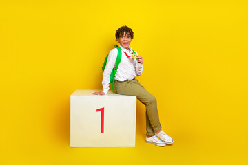 Confident schoolboy showing medal while sitting on podium against vibrant yellow background...