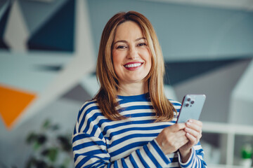 Smiling woman in casual striped outfit holding a smartphone indoors showcasing happiness and leisure