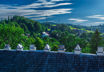 Wooden rooftops with white chimneys overlook a green valley and scattered houses. The view from...