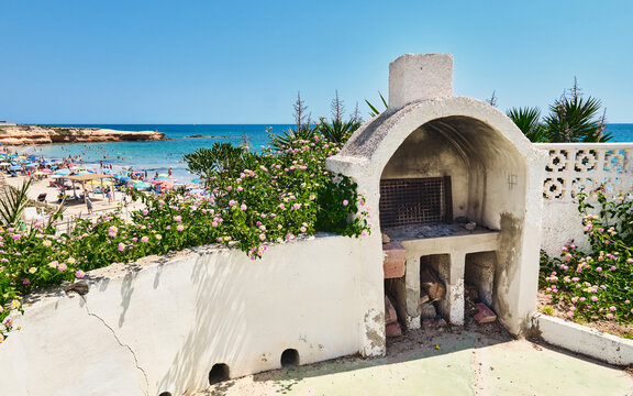 Weathered outdoor barbecue with cracked walls and flowers in the foreground, overlooking a crowded sandy beach with umbrellas and swimmers enjoying a sunny summer day by the sea. Costa Blanca. Spain