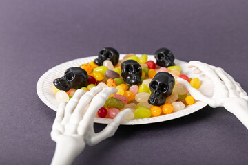 Skeleton hands holding a plate of Halloween candy skulls and jelly beans on a purple background, close-up