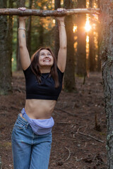 A cheerful young woman in the forest exercising by pulling herself up on a tree branch like on a horizontal bar. Concept of strength, energy, outdoor fitness, and harmony with nature.