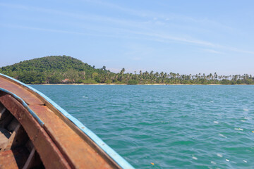 Obraz premium A wooden boat glides through clear turquoise waters, approaching a lush tropical island with palm trees and sandy beaches under a bright blue sky.