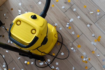 Bright yellow vacuum cleaner on wooden floor covered with scattered colorful star confetti after party celebration cleanup. Concept of household maintenance, cleaning services and post-event tidying.