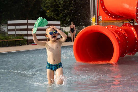 Young boy crying after accident at water park with green cap in hand standing near red tube slides in shallow pool. Concept of water safety, child protection and aquatic playground security measures.