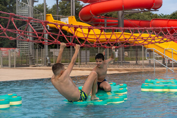 Happy boys playing in outdoor water park pool, climbing on floating platforms and holding red rope net with colorful slides in background on summer day. Concept of fun, active lifestyle and adventure.