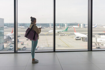 Fototapeta premium A traveler stands by a large window in an airport terminal, looking out at various airplanes on the tarmac. She holds a mobile device and appears to be waiting for her flight.