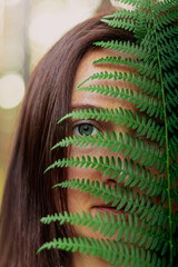 Close-up of a young woman’s face seen through the slits of a fern leaf, creating a natural frame...