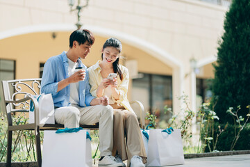 A young couple sits on a bench at an outdoor shopping mall, smiling while looking at a smartphone. Shopping bags around them symbolize modern consumer lifestyle and digital connection
