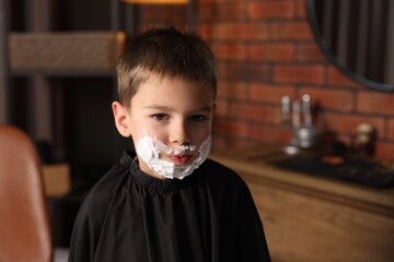 Little boy with shaving foam on his face in barbershop, space for text