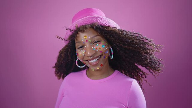 Playful young woman with colorful face jewels and pink hat is smiling and gesturing towards camera against a vibrant pink background, creating a cheerful and expressive mood	