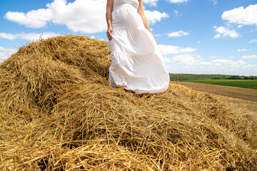 Only legs of a beautiful girl walking on a haystack of straw in a wheat field in a white dress are...