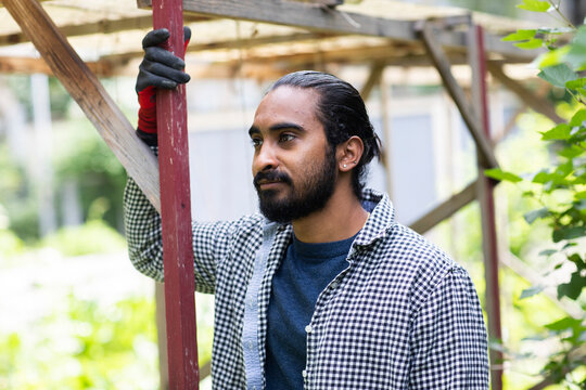 Man in checkered shirt and gloves stands thoughtfully outdoors by wooden structure. Germany