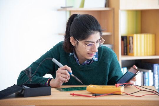 Young woman using a soldering iron and multimeter at a desk with bookshelves in the background. Germany