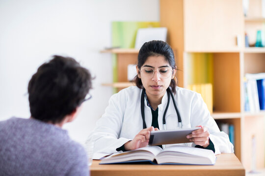 Doctor consulting patient at desk with books and tablet in bright modern office. Germany