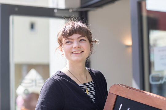 Smiling young woman holding a chalkboard sign outside a modern building. Germany