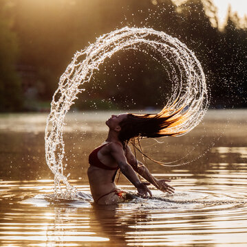 Woman in bikini flips wet hair creating an arc of water in a sunlit lake. WA, USA